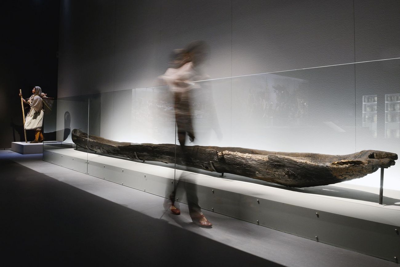 Ausstellungsraum mit einem langen, alten Einbaumboot hinter Glas, während eine Person unscharf daran vorbeigeht.