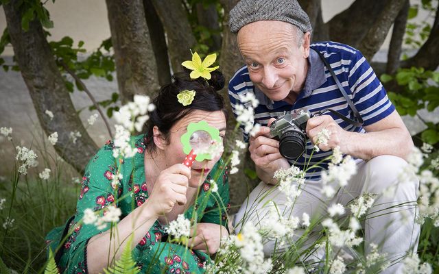 Zwei Personen knien in einer Wiese mit weissen Blüten und betrachten aufmerksam etwas im Gras, während eine der Personen eine Kamera in den Händen hält. Beide tragen farbenfrohe Kleidung und befinden sich unter einem Baum mit dichtem Stammwerk.