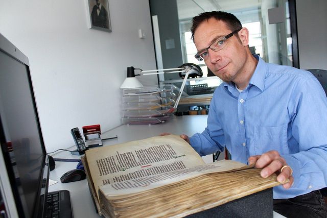 Eine Person sitzt am Schreibtisch und betrachtet ein altes, aufgeschlagenes Buch.