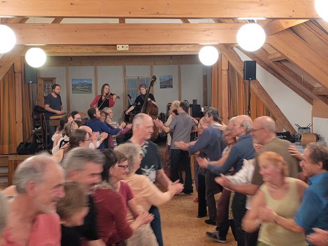 In einem Holzsaal tanzen zahlreiche Menschen gemeinsam zu Live-Musik einer kleinen Band. Die lebhafte Szene vermittelt Gemeinschaft, Bewegung und festliche Stimmung.