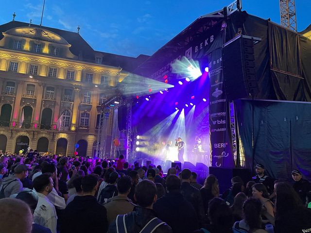 Une scène avec de la musique live illumine la soirée, tandis qu'une grande foule est rassemblée devant. La scène montre un concert animé dans un décor urbain historique avec une ambiance lumineuse intense.