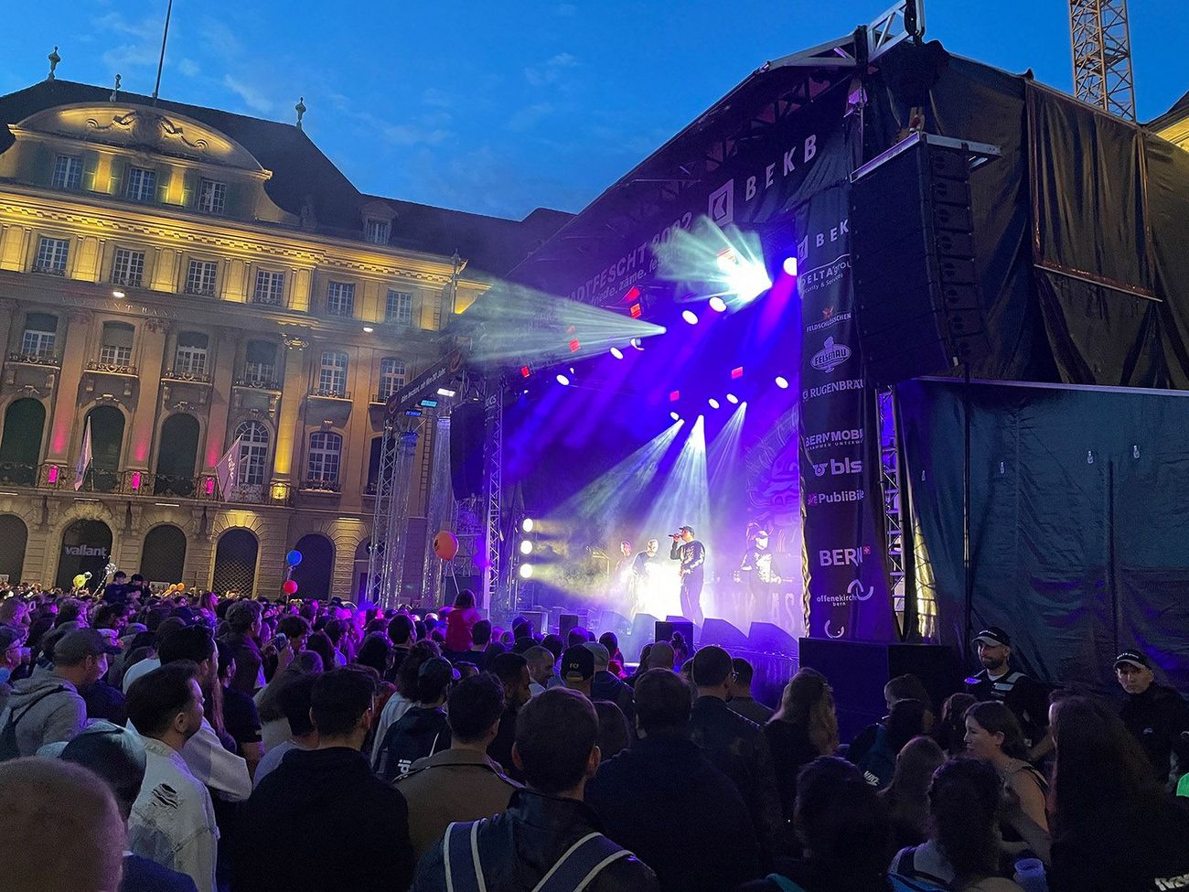 Eine Bühne mit Live-Musik beleuchtet den Abend, während eine grosse Menschenmenge davor versammelt ist. Die Szene zeigt ein lebendiges Konzert in einer historischen Stadtkulisse mit intensiver Lichtstimmung.