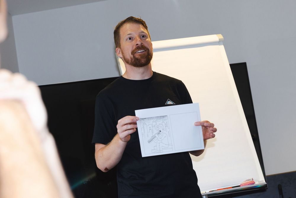 Eine Person steht vor einem Flipchart und präsentiert eine Skizze auf einem Blatt Papier. Die Szene zeigt eine kreative Vorstellung oder Ideation in einem Workshop- oder Präsentationssetting.