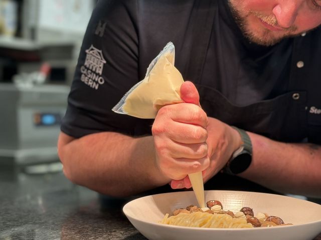 Eine Person richtet ein Gericht an und drückt mit einer Spritztüte sorgfältig eine Creme oder Sauce auf die Speisen. Die Nahaufnahme zeigt präzise Küchenarbeit und detailorientiertes Anrichten.