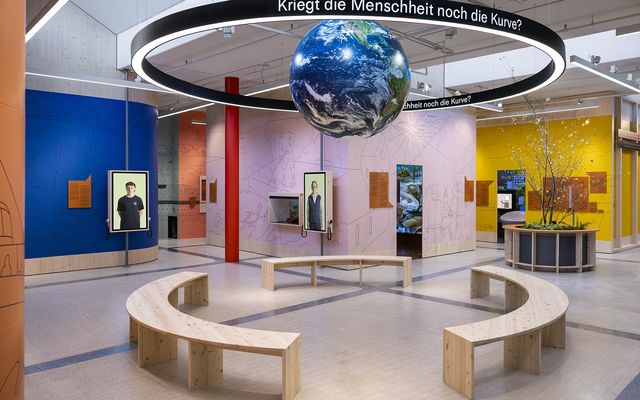Moderne Ausstellung mit hängender Erdkugel, ringförmiger Leuchte, bunten Wänden und Holzsitzbänken.