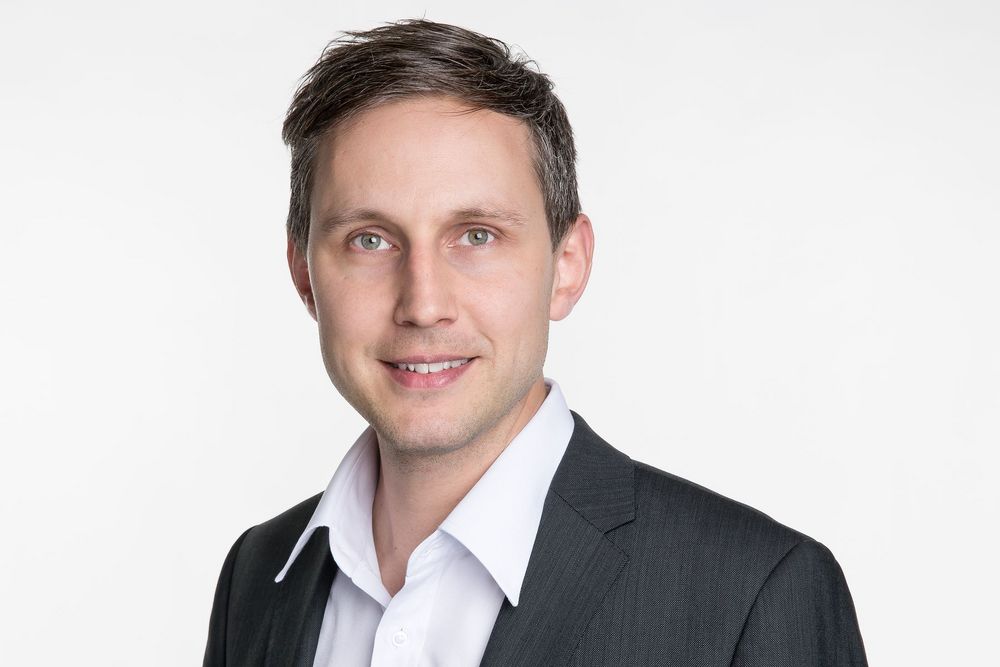Mann in dunklem Anzug und weissem Hemd vor hellem Hintergrund.