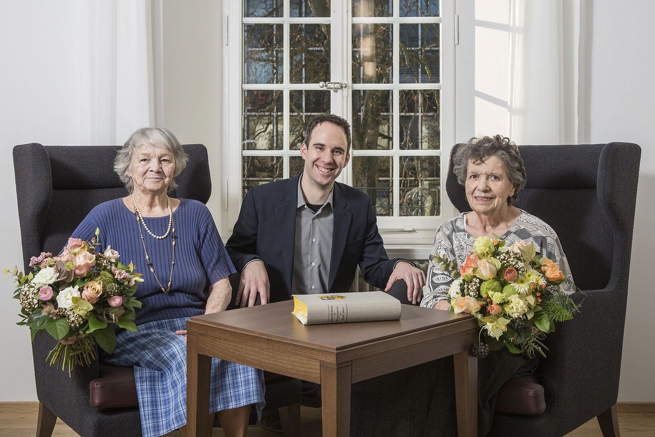 Zwei ältere Frauen mit Blumensträussen sitzen mit einem jüngeren Mann an einem kleinen Tisch.