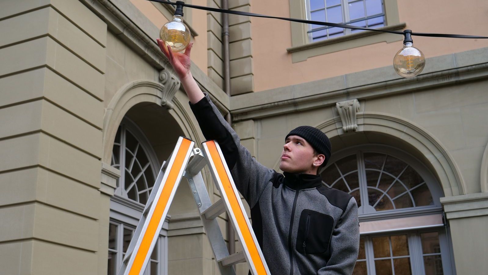 Un apprenti du service de technique du bâtiment monte sur une échelle et accroche une ampoule.