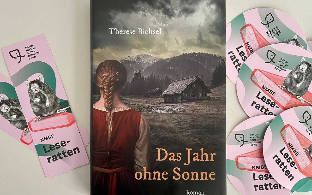 Ein Buch mit dem Titel „Das Jahr ohne Sonne“ liegt auf einem Tisch, flankiert von mehreren farbigen Lesezeichen und Stickern mit dem Motiv einer lesenden Ratte. Das Buchcover zeigt eine Person in historischer Kleidung, die auf eine Berglandschaft mit einem abgelegenen Haus blickt.