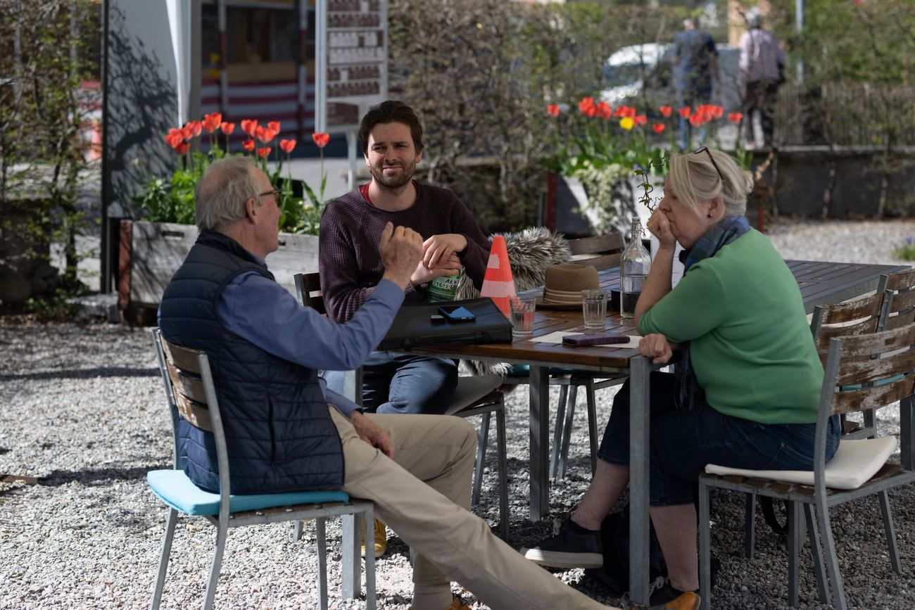 Drei Personen sitzen draussen an einem Tisch und führen ein Gespräch in entspannter Atmosphäre. Die Szene zeigt Austausch und Begegnung in einem grünen, offenen Umfeld.