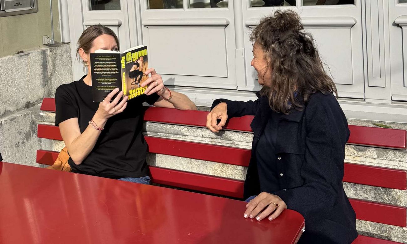 Zwei Frauen im Gespräch auf einer roten Sitzbank, eine liest aus einem Buch vor.