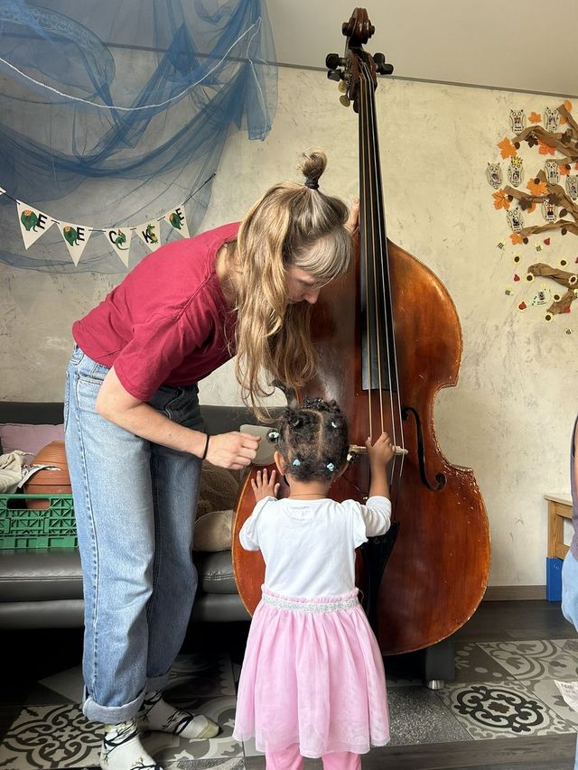 Eine erwachsene Person zeigt einem Kind, wie ein grosses Streichinstrument gespielt wird. Die Szene zeigt gemeinsames Lernen und musikalisches Entdecken in einem gemütlichen Innenraum.