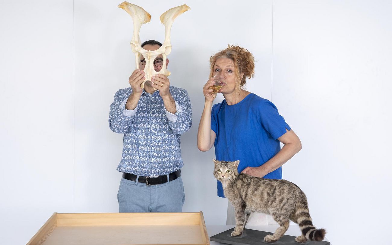 Zwei Personen stehen vor einem Tisch; eine Person hält eine grosse, helle Tiermaske vor das Gesicht, während die andere eine kleinere Tierfigur neben sich hält. Auf dem Tisch steht zudem ein Modell eines gefleckten, katzenartigen Tiers.