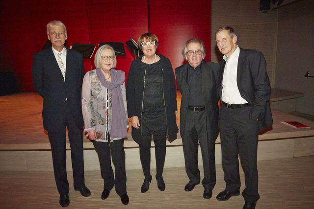 Fünf festlich gekleidete Personen stehen gemeinsam in einem Saal mit roter Wand und Bühne.