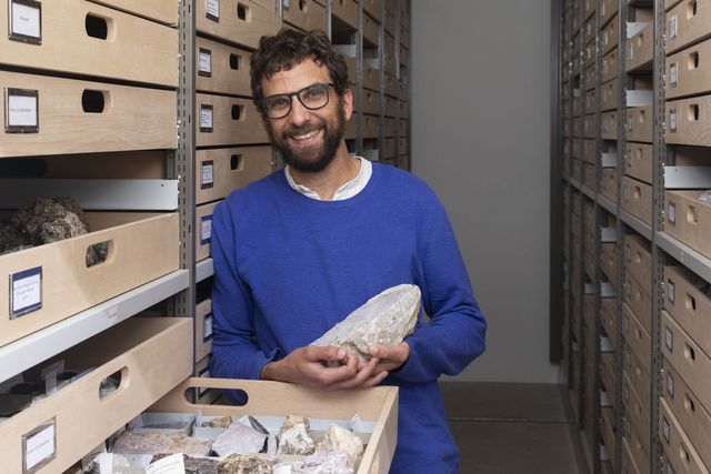 Une personne se tient dans une salle d'archives, entre des étagères remplies de tiroirs contenant des échantillons de roches, et tient dans ses mains un gros minéral. La scène illustre la collecte dans les sciences et la conservation minutieuse de spécimens naturels.