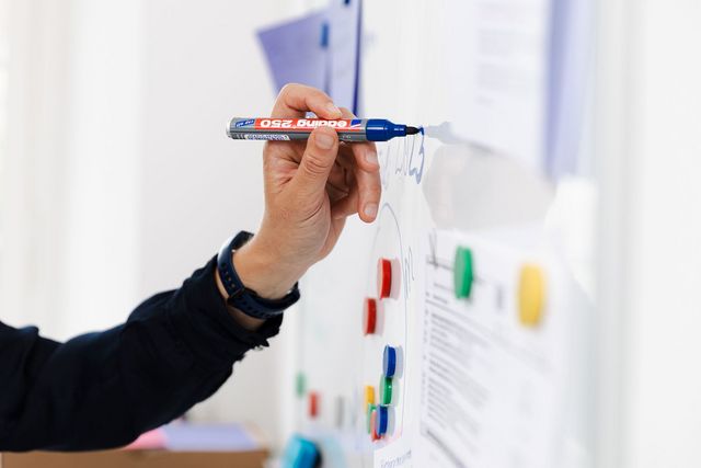 Une main écrit avec un stylo bleu sur un tableau blanc.