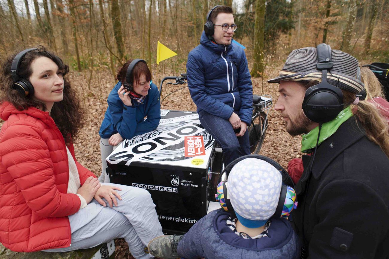 Menschen sitzen im Wlad mit Kopfhörern um eine Soundbox
