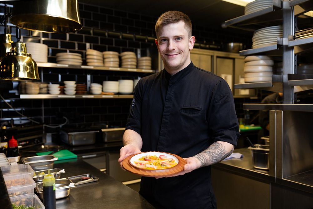 Ein Koch präsentiert ein liebevoll angerichtetes Gericht in einer modernen Restaurantküche. Die Szene zeigt professionelle Zubereitung und kreative Küche auf hohem Niveau.