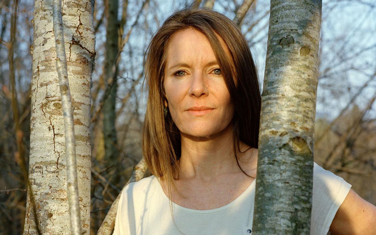 Eine Frau steht zwischen mehreren Baumstämmen in einem Waldgebiet. Die Frau trägt ein helles Oberteil und lehnt leicht an einem der Bäume.