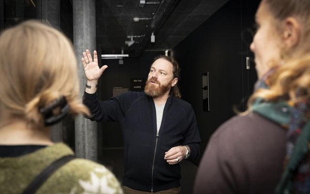 Person erklärt einer kleinen Gruppe etwas in einem Museumsraum und gestikuliert mit erhobener Hand.