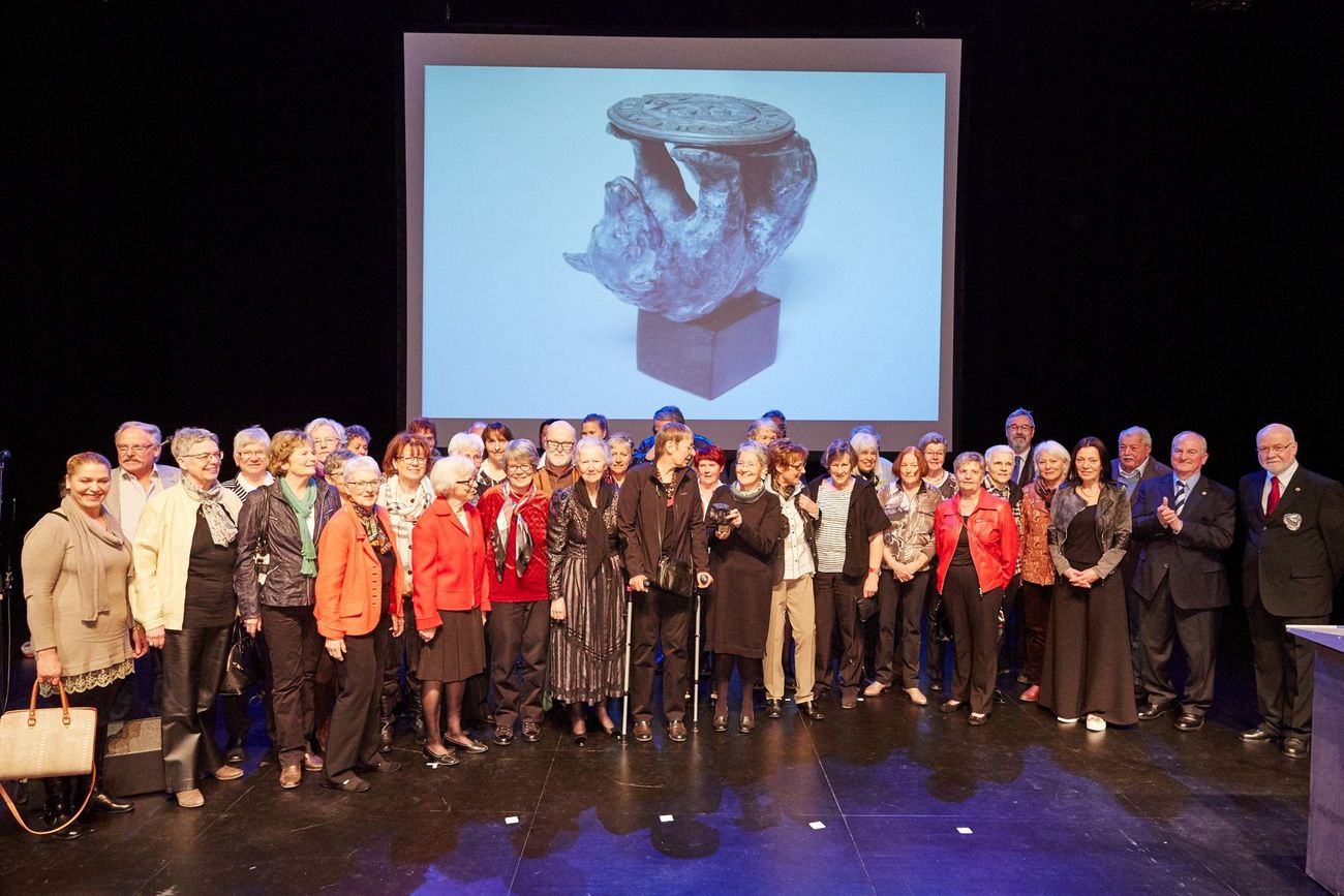 Eine grosse Gruppe Menschen steht auf einer Bühne vor einer Leinwand mit einer Skulpturprojektion