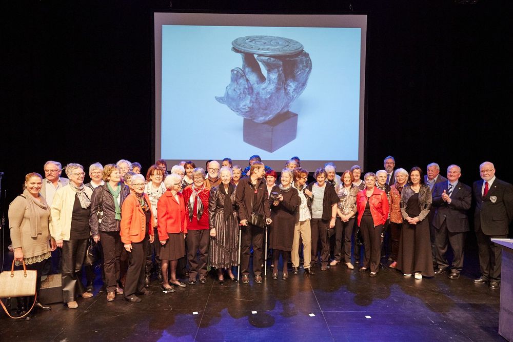 Eine grosse Gruppe Menschen steht auf einer Bühne vor einer Leinwand mit einer Skulpturprojektion
