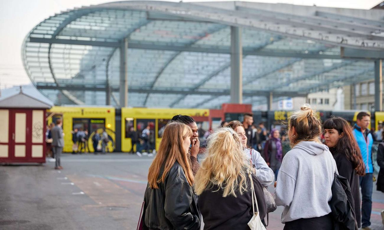 Eine Gruppe von Menschen steht vor dem Bahnhof und unterhält sich, während im Hintergrund gelbe Trams und eine grosse Glasüberdachung zu sehen sind.