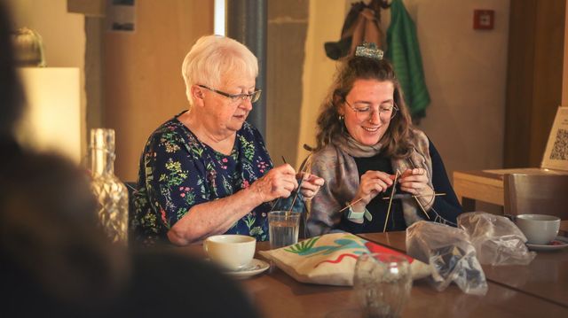 Zwei Personen sitzen an einem Tisch und stricken gemeinsam mit Nadeln und Wolle. Die Szene zeigt gemeinsames Handarbeiten und den Austausch zwischen Generationen in einer gemütlichen Umgebung.
