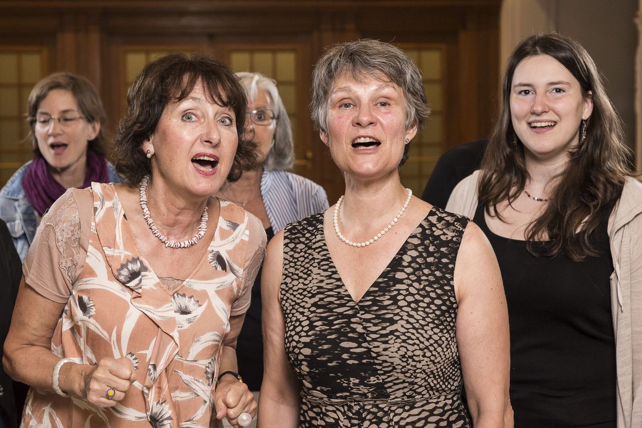Mehrere Personen stehen dicht beisammen in einem Innenraum; zwei Frauen im Vordergrund tragen elegante Kleidung und Schmuck.