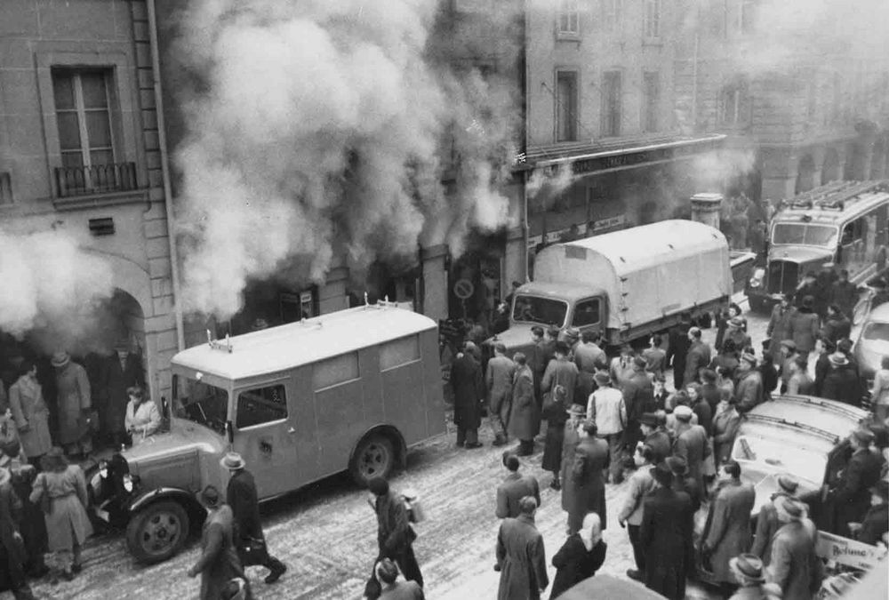 Alte Schwarz‑Weiss‑Aufnahme: Menschen beobachten Feuerwehrfahrzeuge und starken Rauch, der aus einem Gebäude dringt.