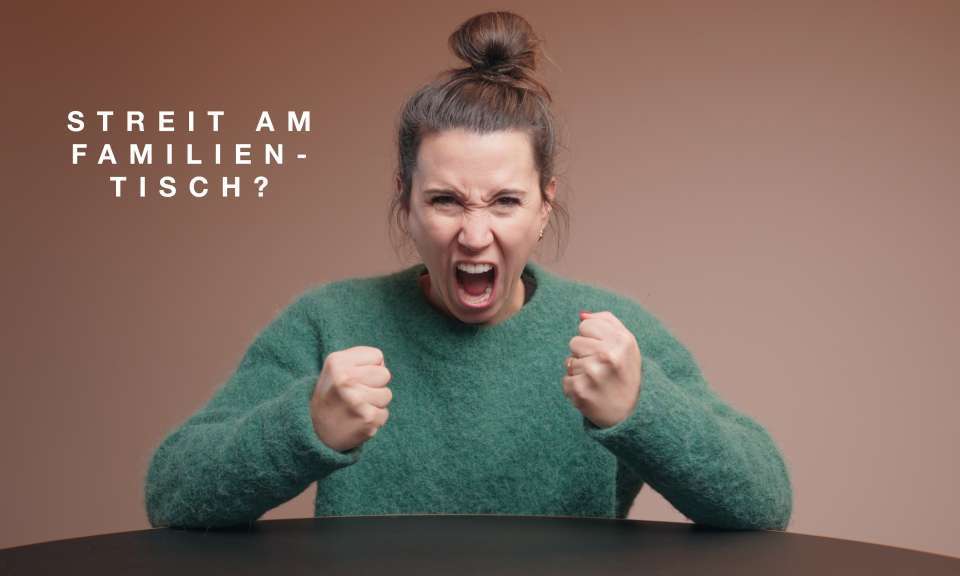 Eine Person sitzt an einem Tisch und hält beide Hände zu Fäusten vor dem Körper, als würde eine angespannte Situation dargestellt. Links im Bild steht der eingeblendete Text „STREIT AM FAMILIEN­TISCH?“.