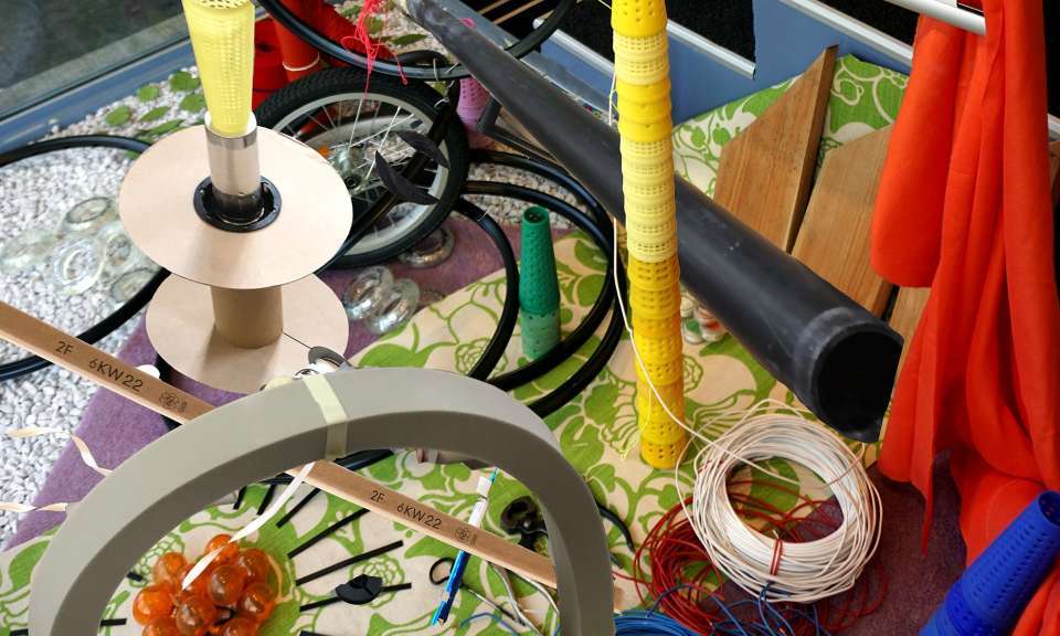Ein farbenfroher Tisch voller Materialien wie Kunststoffrohre, Holzlatten, Fahrradräder, Garnspulen, Kabel und Stoffstücke liegt chaotisch arrangiert nebeneinander. Die vielfältigen Gegenstände wirken wie eine Sammlung von Rest- und Bastelmaterialien für kreative Projekte.