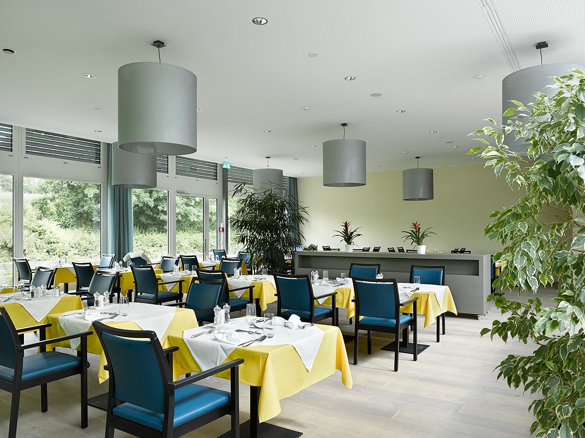 Helles Speiserestaurant im Burgerspittel Viererfeld mit gelb gedeckten Tischen, blauen Stühlen und grossen runden Hängelampen.
