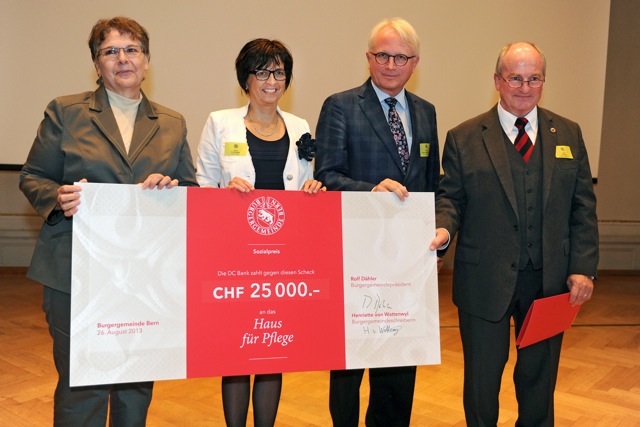 Vier Personen stehen in einem Saal und halten gemeinsam einen grossen Spendenscheck über 25’000 CHF mit der Aufschrift ‘Haus für Pflege’. Eine Person daneben hält einen roten Umschlag