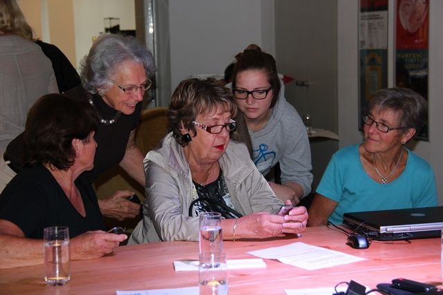 Jugendliche unterstützen ältere Menschen am Tisch beim Umgang mit Smartphone und Laptop.
