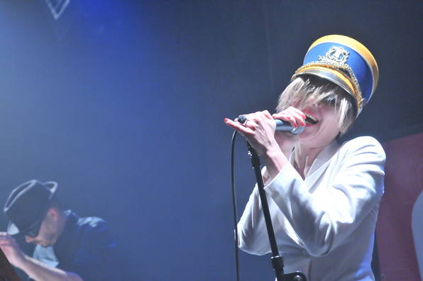 Sängerin mit uniformeller Mütze performt auf der Bühne am Mikrofon, begleitet von einem Musiker im Hintergrund.